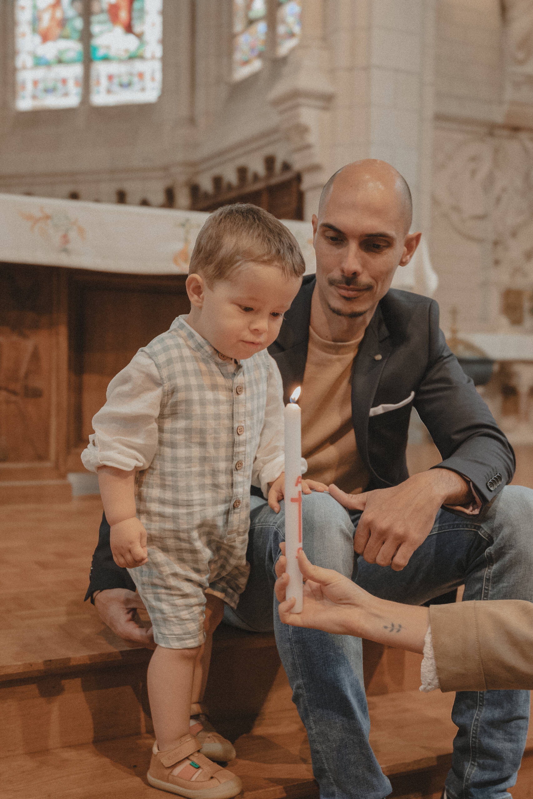 Enfant soufflant sur la flamme du cierge pascal, accompagné de son père assis sur les marches devant l’autel de l’église de L’Herbergement en Vendée, lors de son baptême. Image réalisée par Solène photographe.
