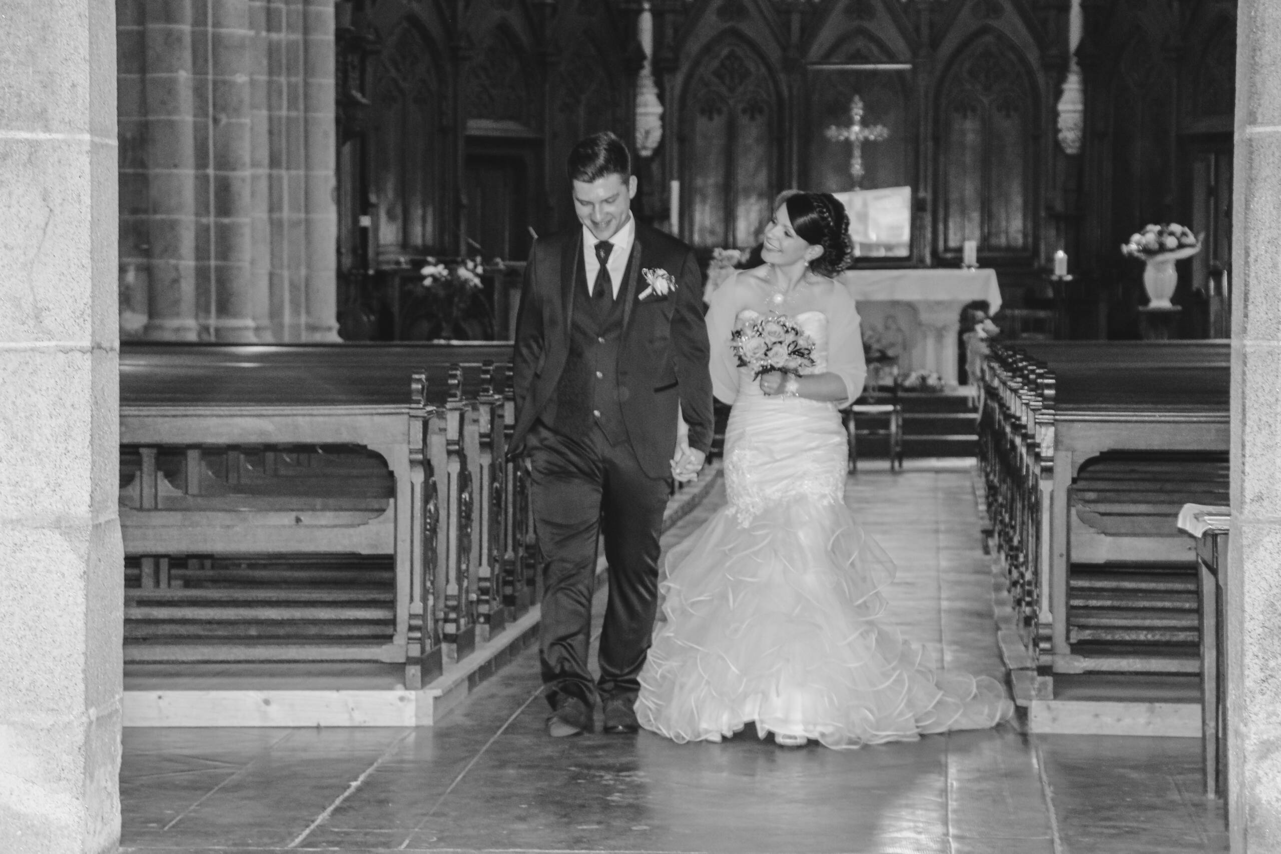 Mariés se tenant la main en sortant de l’église après leur cérémonie religieuse pendant leur mariage, réalisée en noir et blanc par Solène photographe.