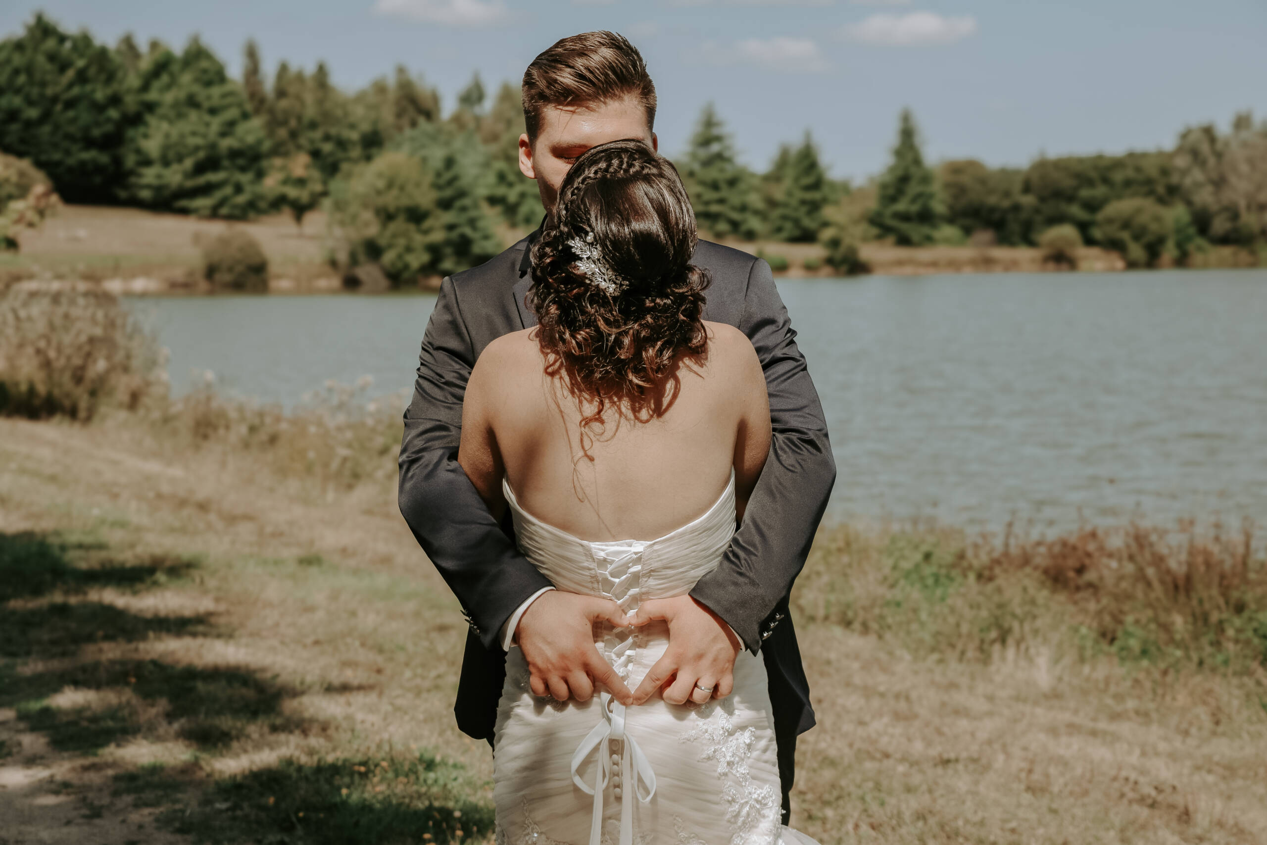 Couple de mariés enlacé, le marié forme un cœur avec ses doigts dans le dos de sa femme, lors des photos de couple pendant leur mariage, réalisée par Solène photographe.