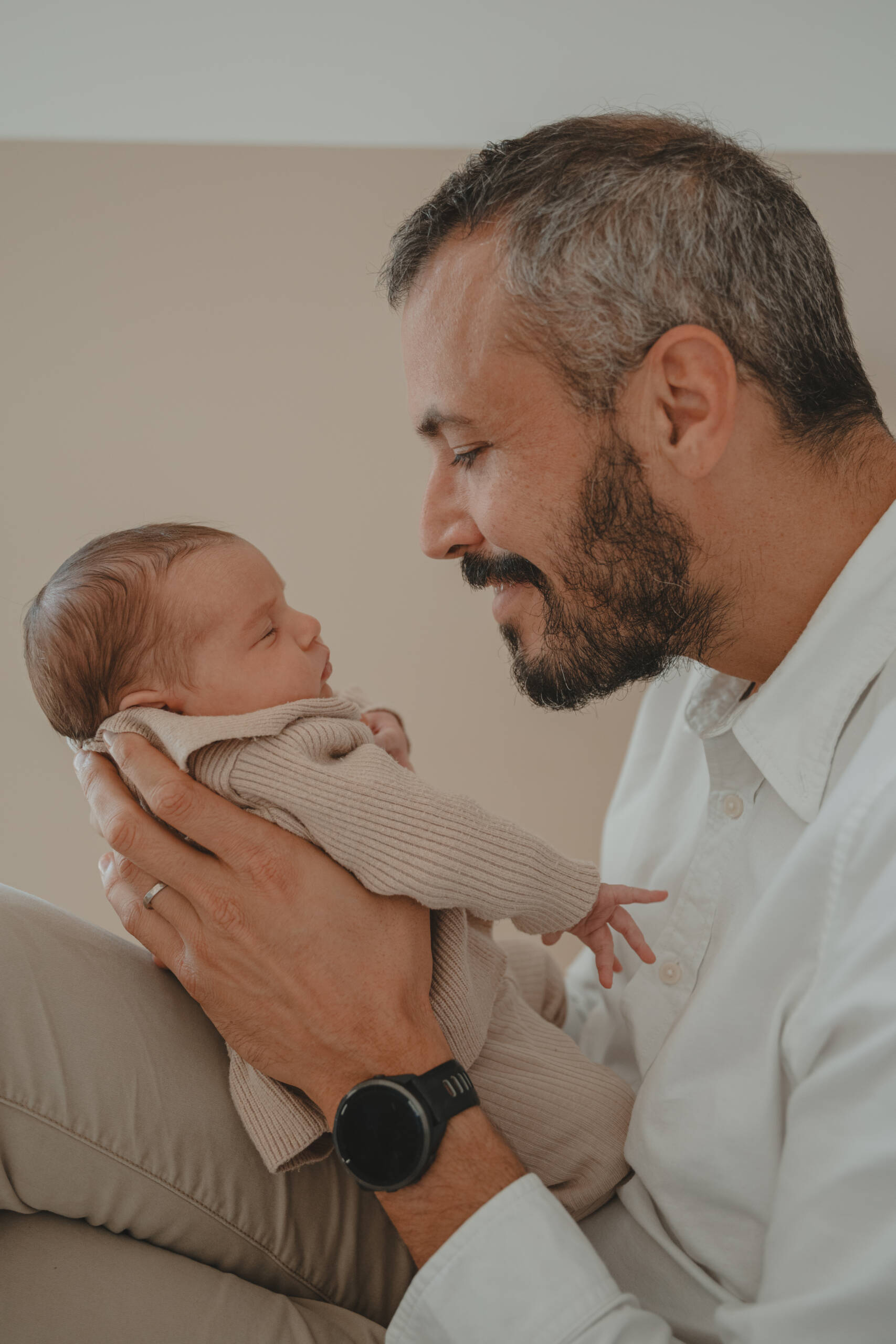 Père assis tenant son nourrisson dans ses mains face à face l'un de l'autre, lors d’une séance photo naissance à domicile à La Ferrière en Vendée, réalisée par Solène photographe.