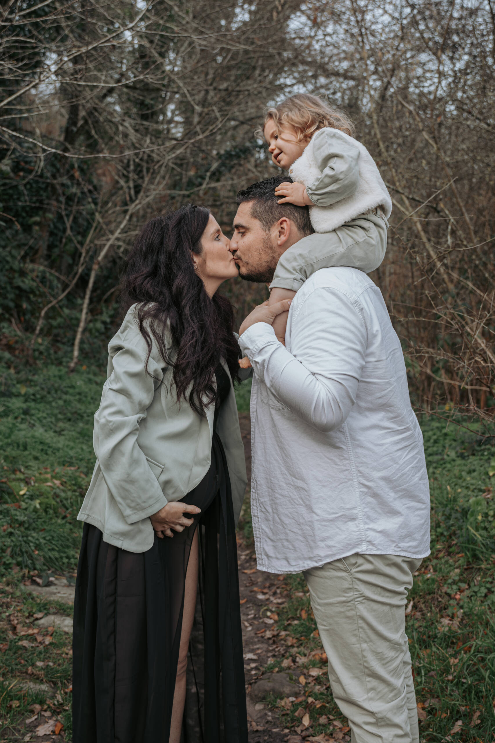 Femme enceinte embrassant son conjoint, qui tient sa fille sur ses épaules, lors d’une séance photo grossesse à Tiffauges en Vendée, réalisée par Solène photographe.