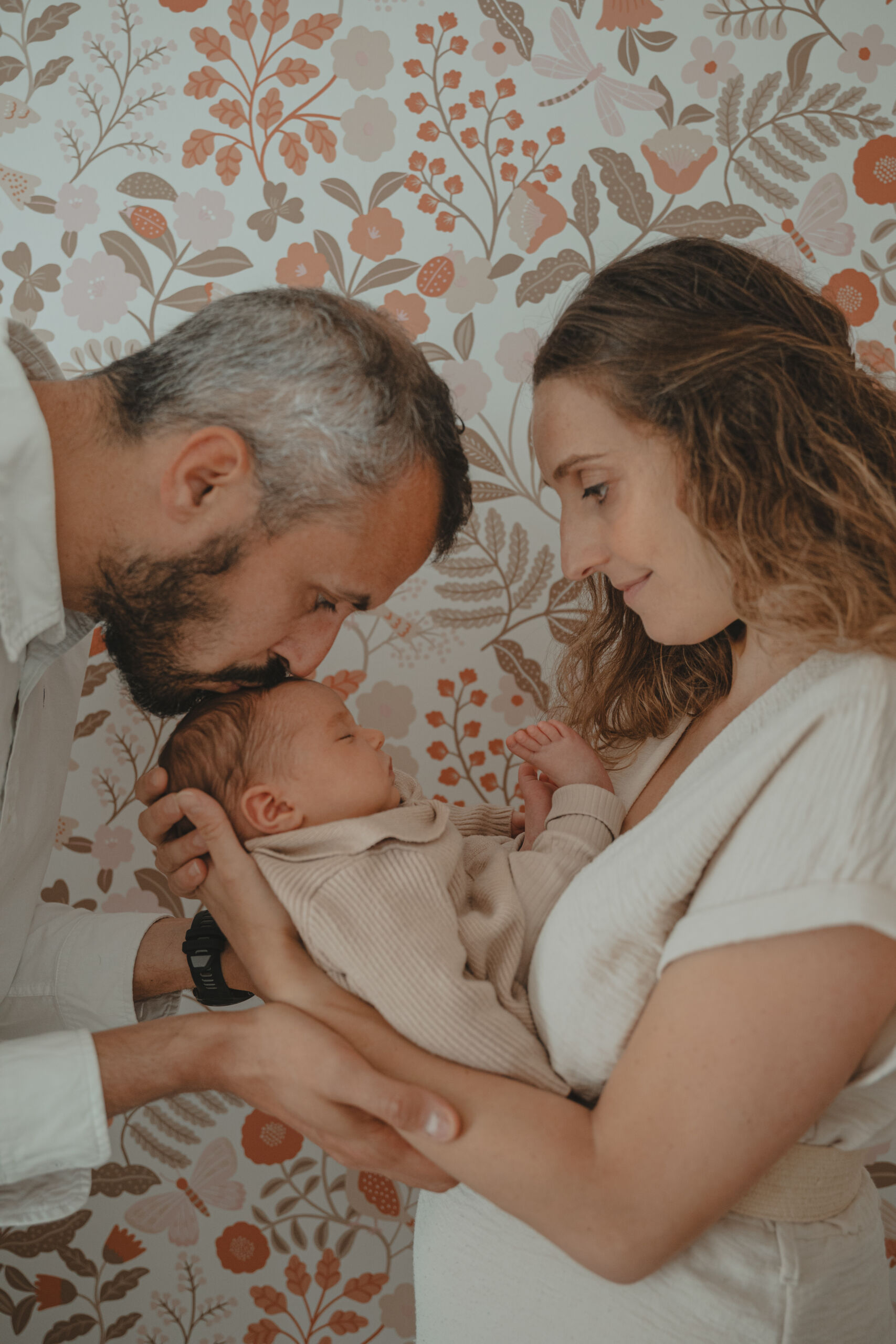 Mère regardant son nourrisson dans ses bras pendant que le papa embrasse le nourrisson sur le front, lors d’une séance photo naissance à domicile à La Ferrière en Vendée, réalisée par Solène photographe.