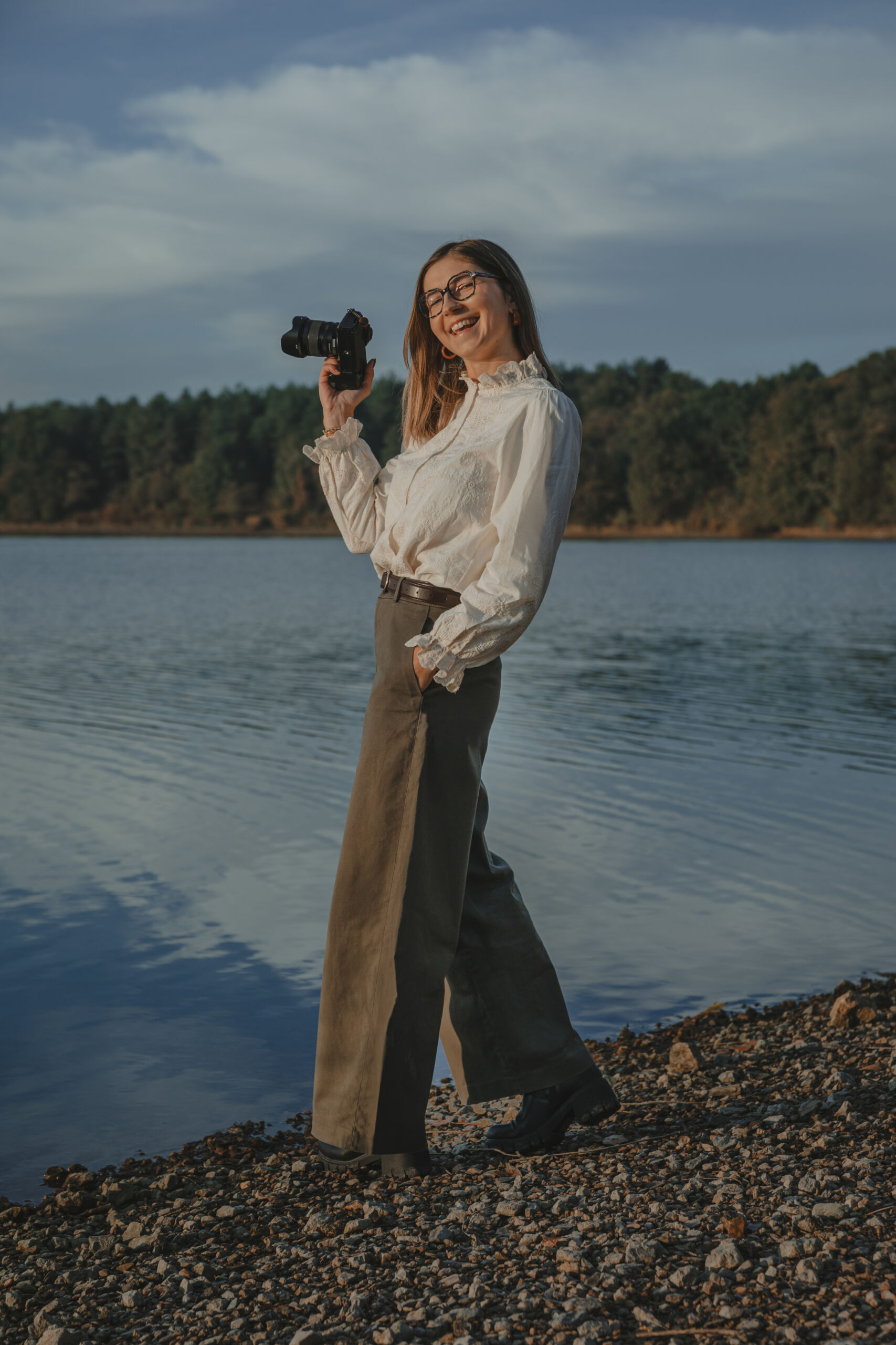 Femme souriante, appareil photo à la main à hauteur de tête, debout près d'un lac, lors d’une séance portrait professionnel à La Roche-sur-Yon en Vendée, réalisée par Solène photographe.