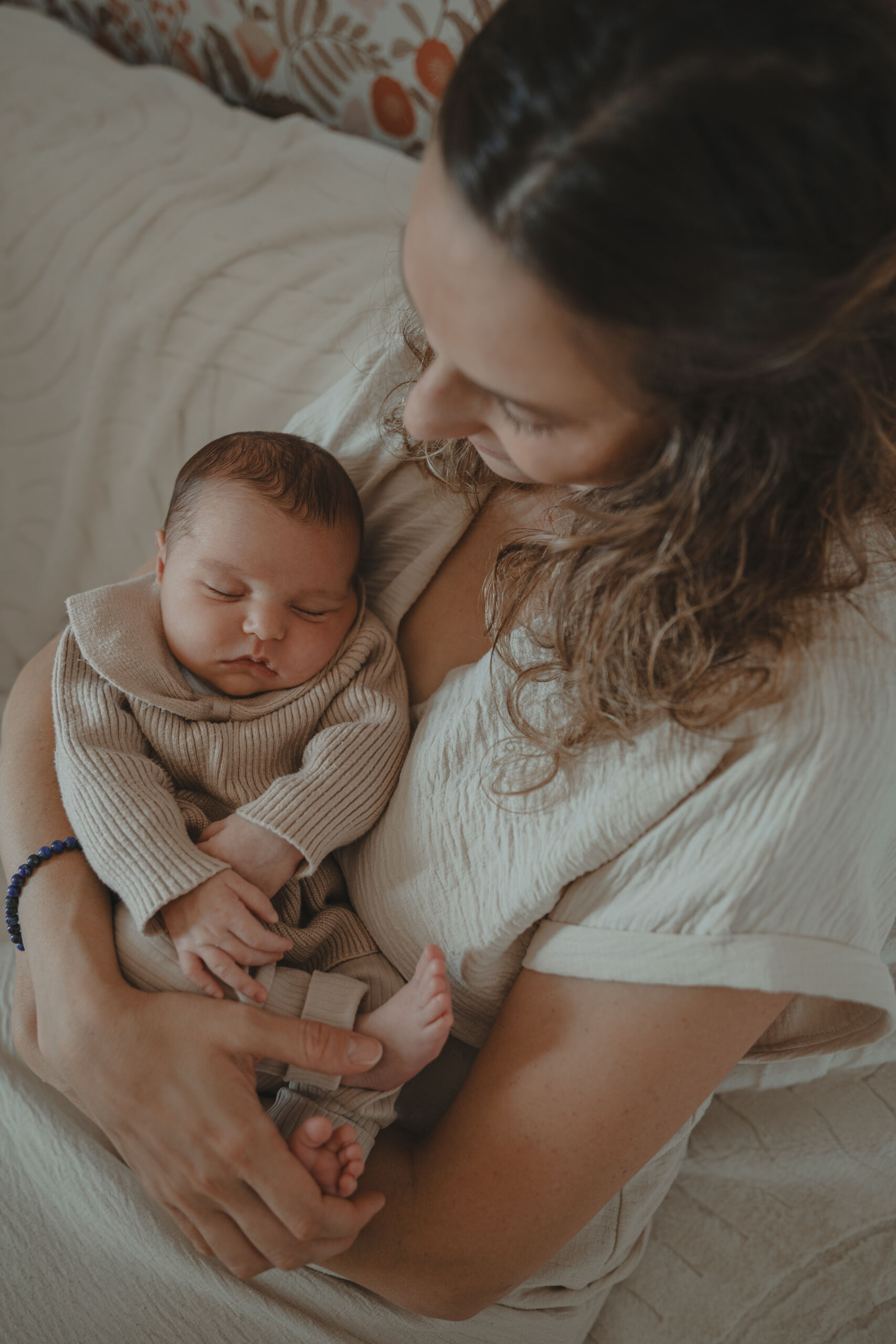 Nourrisson dans les bras de sa mère assise, vue du dessus, lors d’une séance photo naissance à domicile à La Ferrière en Vendée, réalisée par Solène photographe.