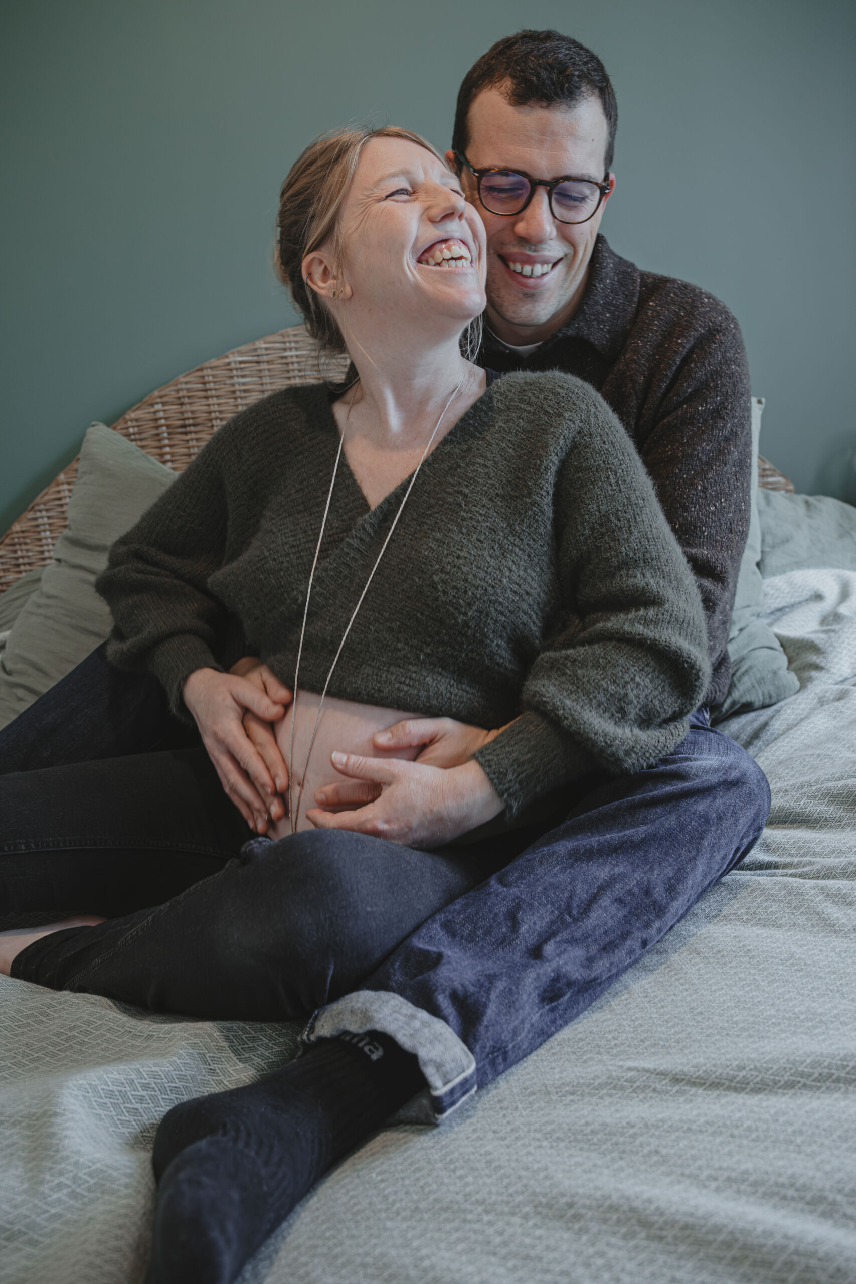 Futurs parents complices et heureux, sur un lit et rejoignant leurs mains sur le ventre rond de la femme, lors d’une séance photo grossesse à Dompierre-sur-Yon en Vendée, réalisée par Solène photographe.