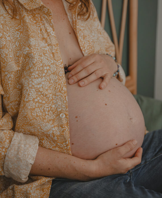 Femme enceinte assise en tailleur sur un lit présentant une main au-dessus et une main en-dessous de son ventre, lors d’un shooting confiance en soi à Montbert en Loire- Atlantique, réalisée par Solène photographe.