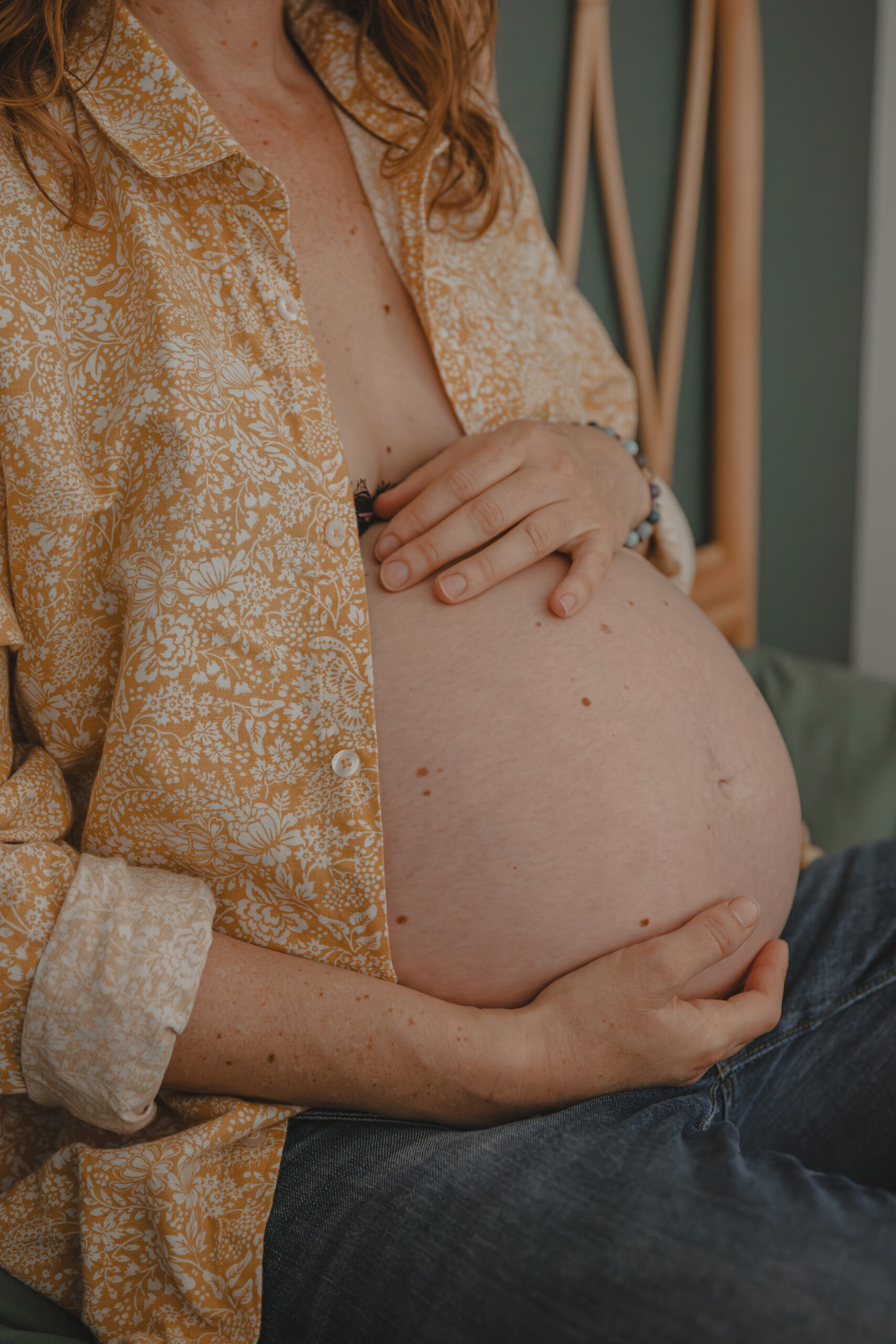 Femme enceinte assise en tailleur sur un lit présentant une main au-dessus et une main en-dessous de son ventre, lors d’un shooting confiance en soi à Montbert en Loire- Atlantique, réalisée par Solène photographe.