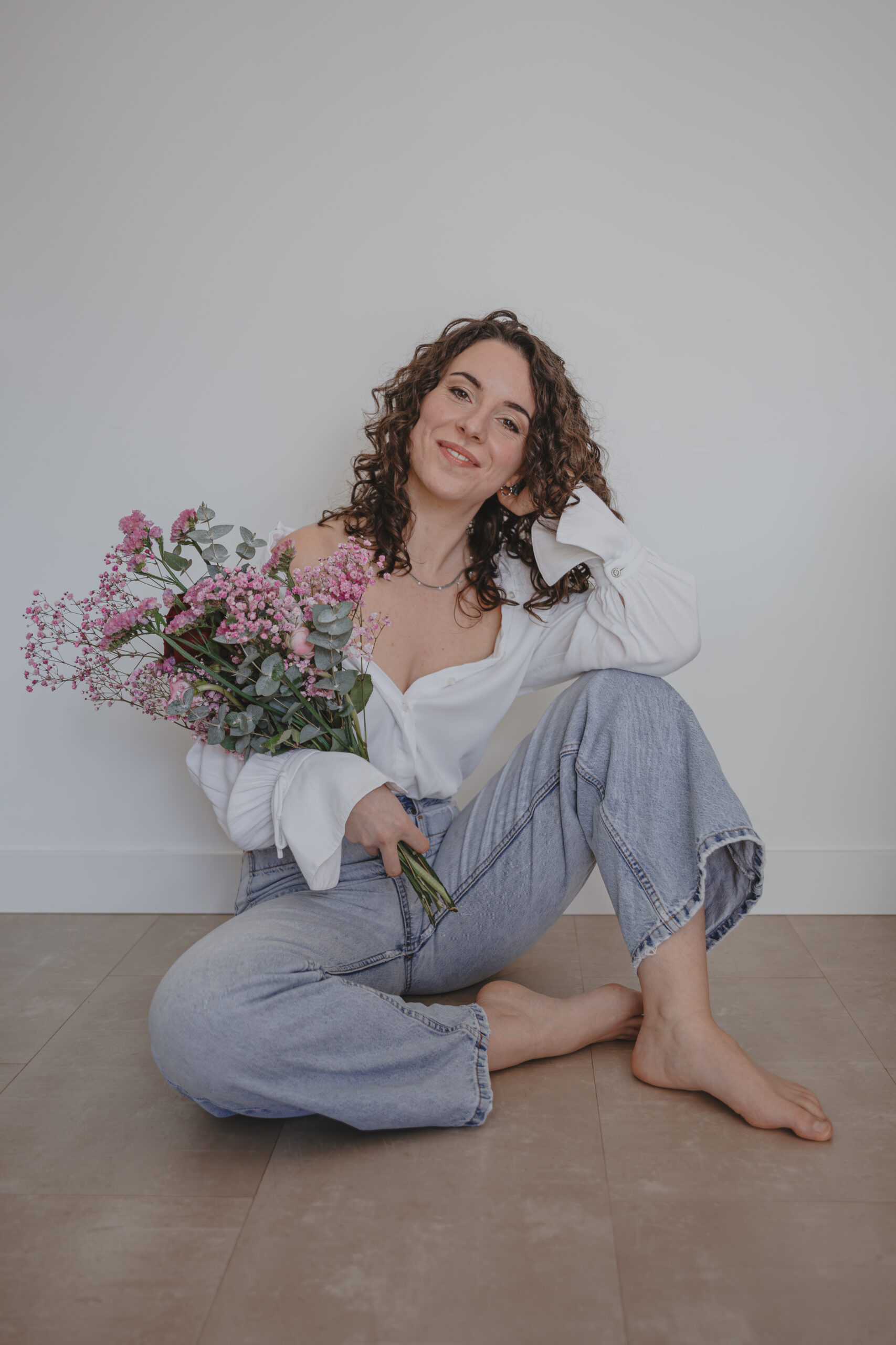 Femme souriante en chemise blanche avec une épaule dénudée assise par terre et tenant un bouquet de fleurs, lors d’un shooting confiance en soi à Montbert en Loire- Atlantique, réalisée par Solène photographe.