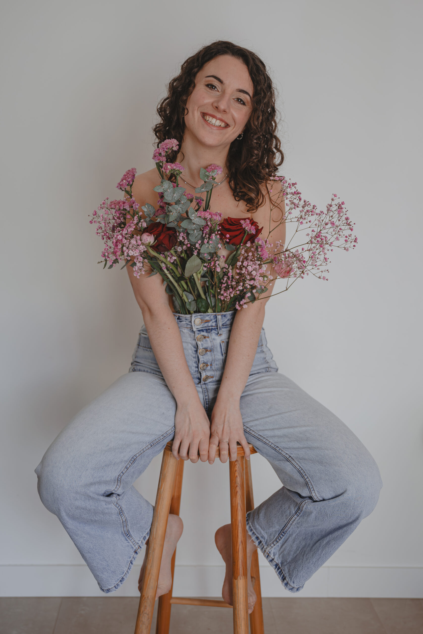 Femme souriante tête penchée assise sur un tabouret en bois et habillée seulement d'un jean où un bouquet de fleurs y dépasse cachant ainsi sa poitrine, lors d’un shooting confiance en soi à Montbert en Loire- Atlantique, réalisée par Solène photographe.