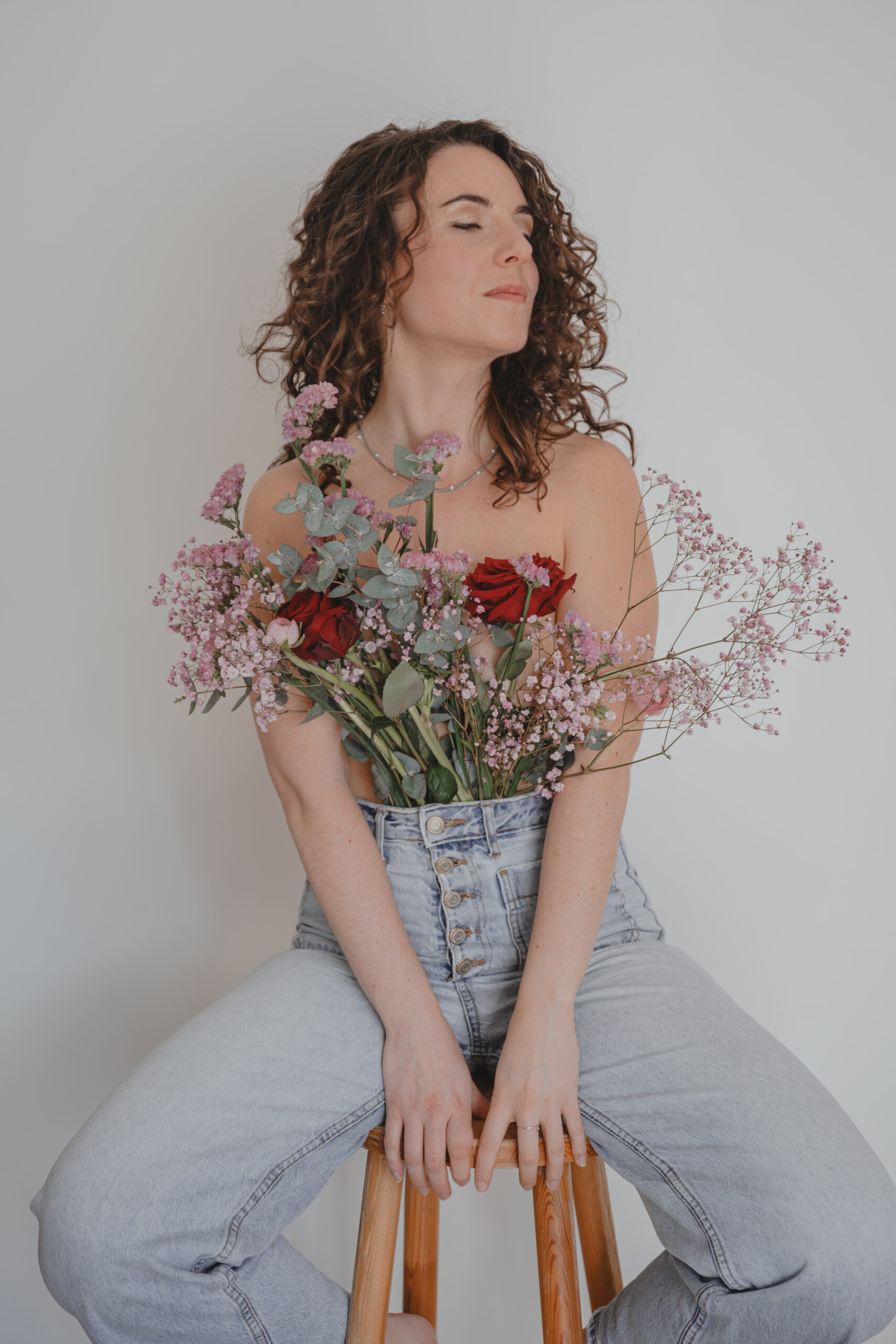 Femme en mouvement assise sur un tabouret en bois habillée d'un jean et d'un bouquet de fleurs cachant ainsi sa poitrine, lors d’un shooting confiance en soi à Montbert en Loire- Atlantique, réalisée par Solène photographe.