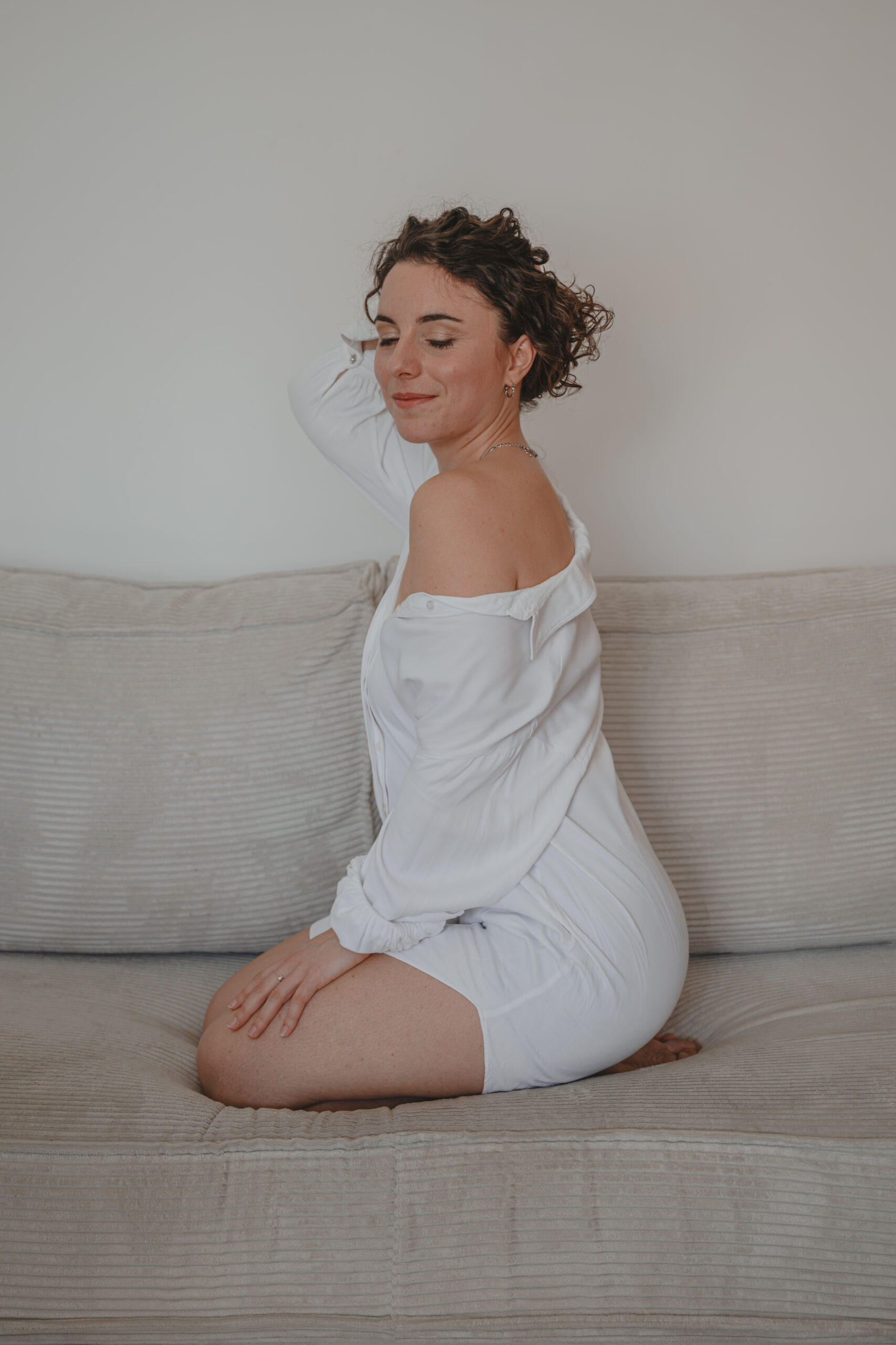 Femme assise sur un canapé habillée d'une chemise blanche avec une épaule dénudée fermant les yeux en se tenant les cheveux, lors d’un shooting confiance en soi à Montbert en Loire- Atlantique, réalisée par Solène photographe.
