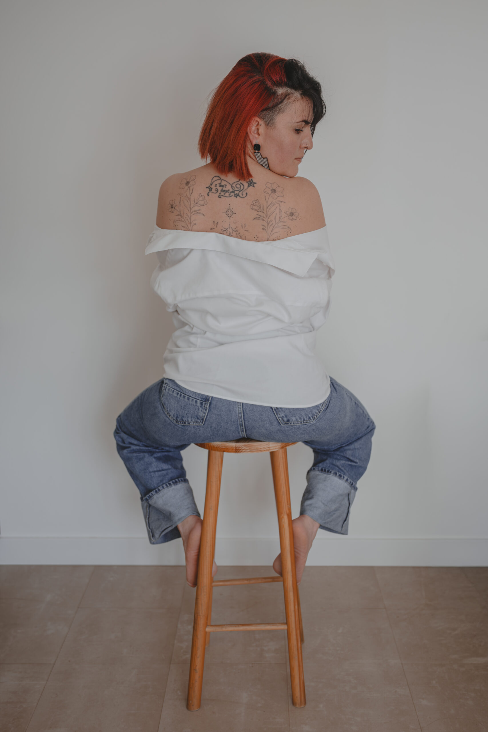 Femme assise sur un tabouret se tenant de dos la tête tournée sur le côté vers le bas et épaules dénudées dévoilant des tatouages, lors d’un shooting confiance en soi à Montbert en Loire- Atlantique, réalisée par Solène photographe.
