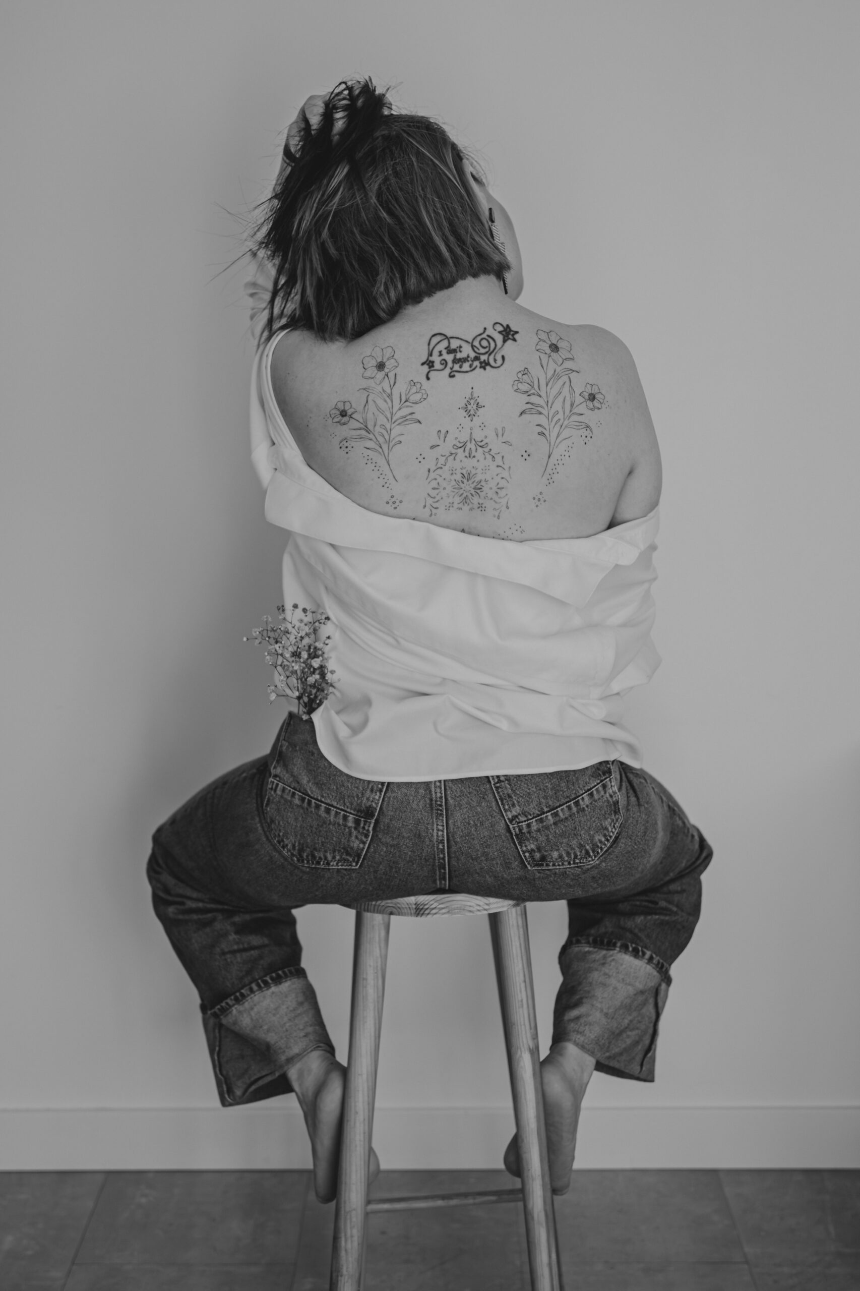 Femme de dos assise sur un tabouret passant les mains dans ses cheveux avec les épaules dénudées dévoilant des tatouages, lors d’un shooting confiance en soi à Montbert en Loire- Atlantique, réalisée en noir et blanc par Solène photographe.