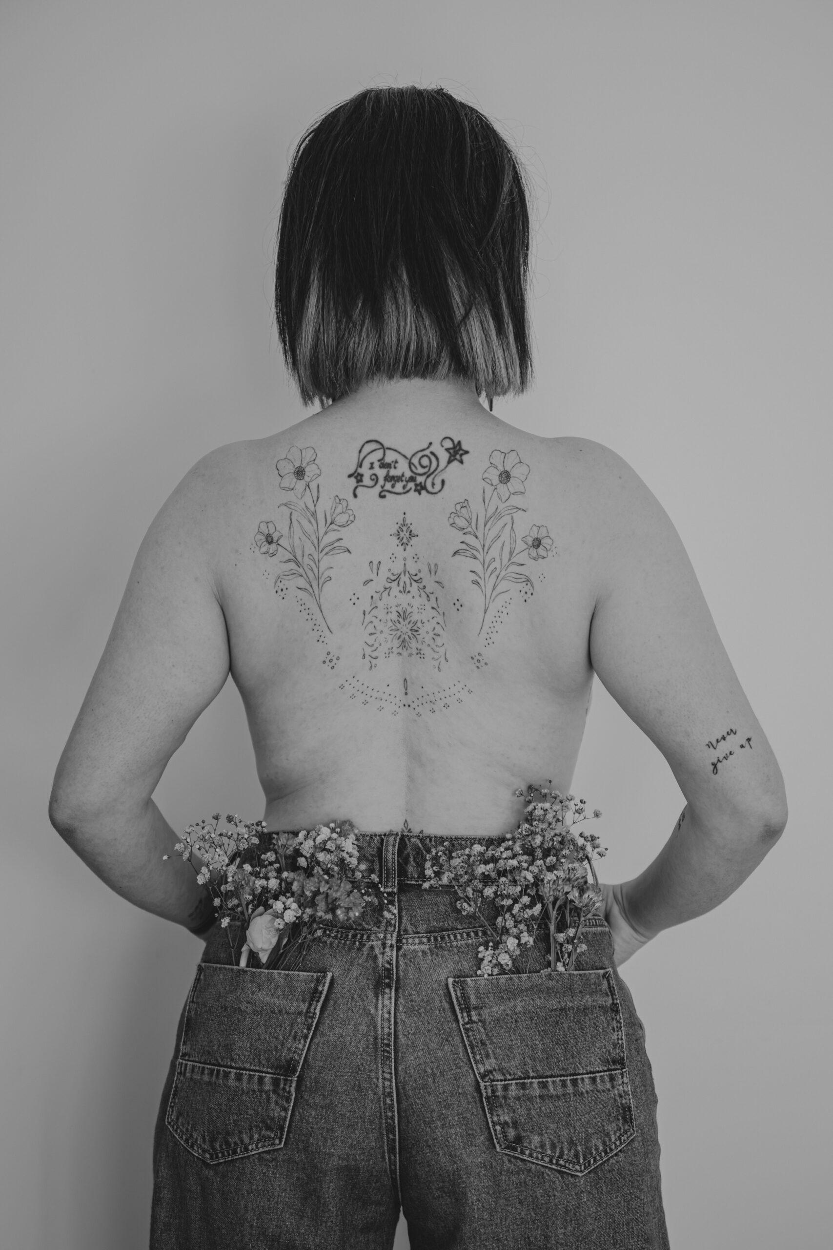 Femme se tenant debout de dos haut nu dévoilant des tatouages posant les mains dans les poches avant et des fleurs dans les poches arrière de son jean, lors d’un shooting confiance en soi à Montbert en Loire- Atlantique, réalisée en noir et blanc par Solène photographe.