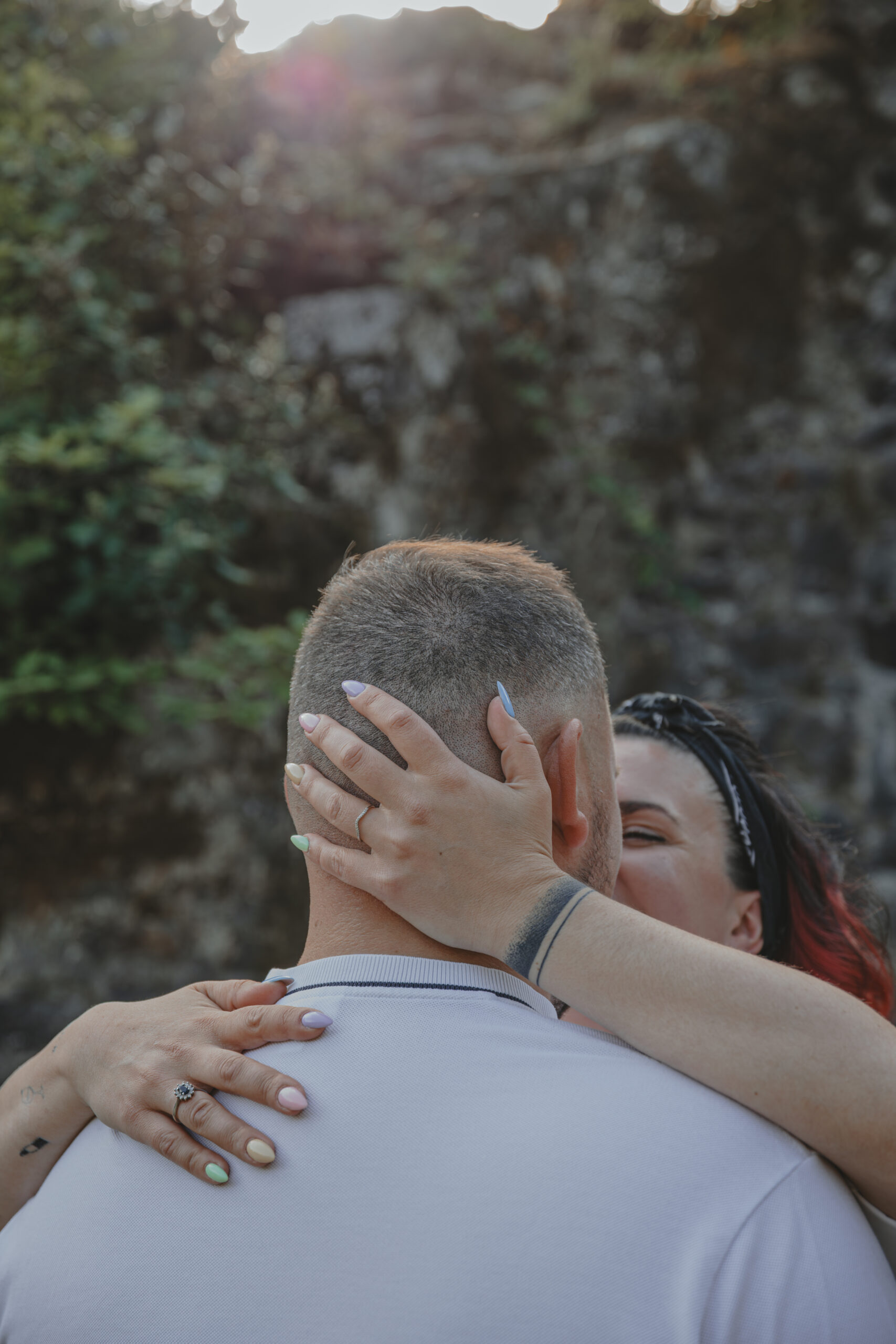 Femme enlaçant son futur mari en posant ses mains sur la nuque et l’épaule de son partenaire, avec une lumière douce en arrière-plan rocheux, mettant en valeur sa bague de fiançailles, lors d’une séance photo engagement au Parc des Remparts à Montaigu-Vendée en Vendée, réalisée par Solène photographe.