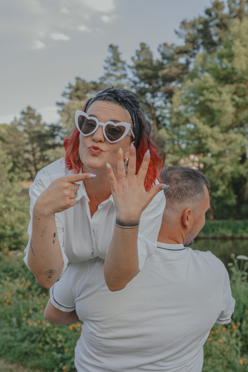 Femme portée sur l'épaule de son futur mari pointant du doigt sa main portant la bague de fiançailles, lors d’une séance photo engagement au Parc des Remparts à Montaigu-Vendée, réalisée par Solène photographe.