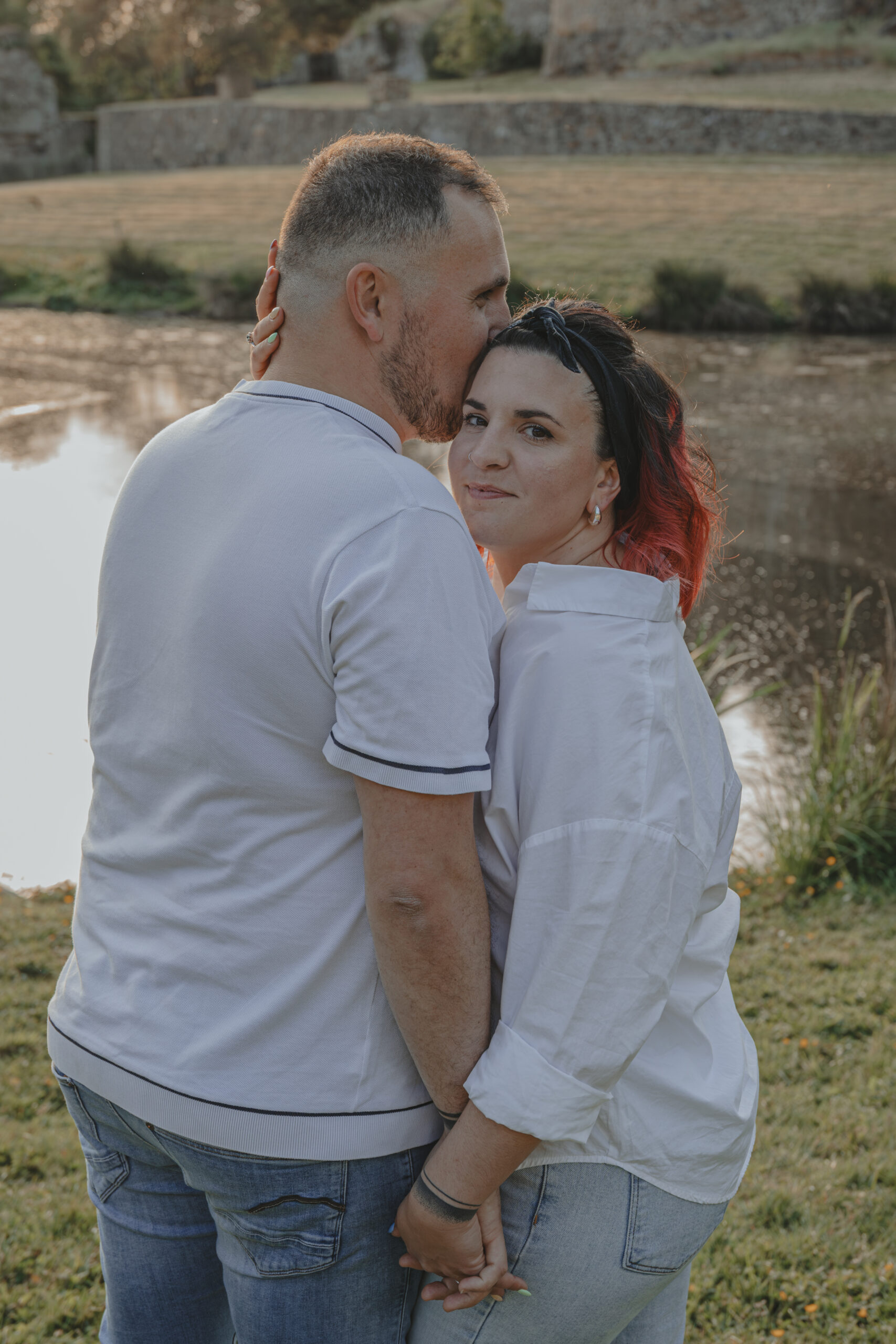 Homme embrassant sur le front sa future femme qui me regarde en se tenant la main, lors d’une séance photo engagement au Parc des Remparts à Montaigu-Vendée, réalisée par Solène photographe.