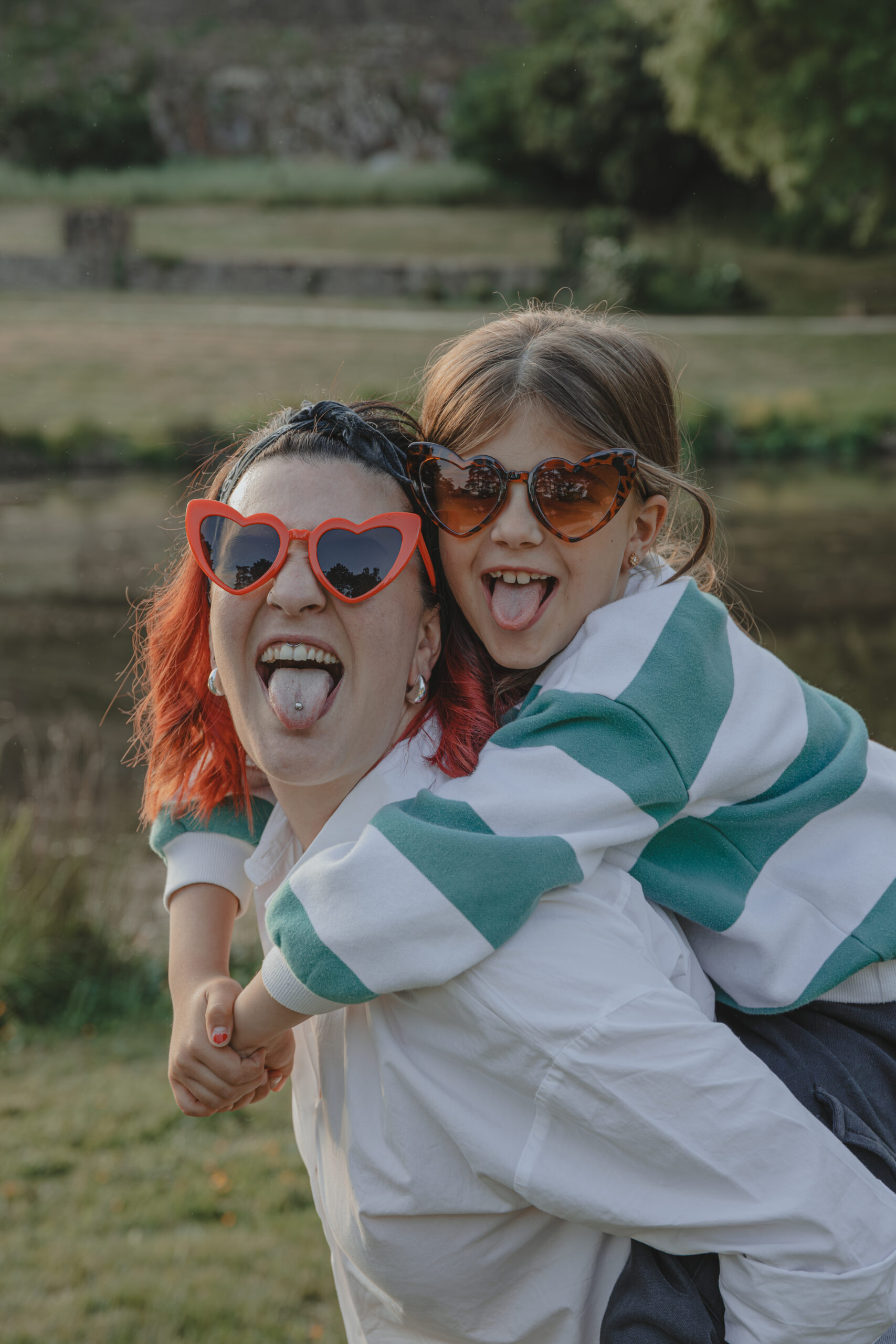 Mère portant sur son dos sa fille et tirant toutes les deux la langue en portant des lunettes cœur, lors d’une séance photo engagement au Parc des Remparts à Montaigu-Vendée, réalisée par Solène photographe.